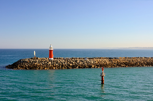 A stone jetty with a red lighthouse at the Fremantle, Australia harbor. People come out to watch the cruise ship come in to port.