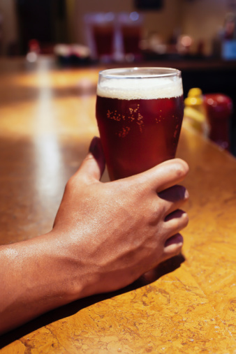 African American person's hand holding a full glass of beer on a bar counter.