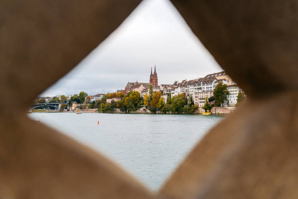 Basel city- A bridge spans Rhein river with a city in the background. The bridge is a stone structure with a red and white sign on it. The city is filled with buildings and a church. The sky is clear and blue