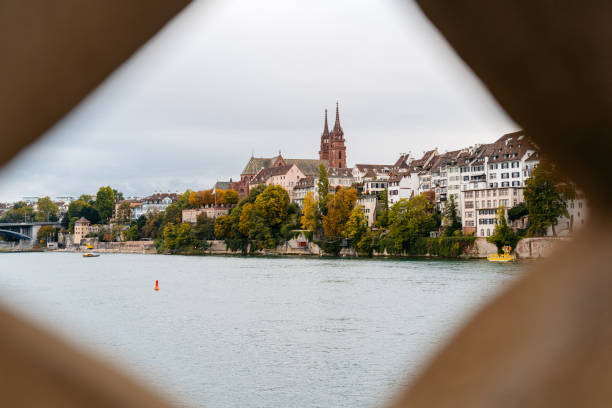 Basel city- A bridge spans Rhein river with a city in the background. The bridge is a stone structure with a red and white sign on it. The city is filled with buildings and a church. The sky is clear and blue