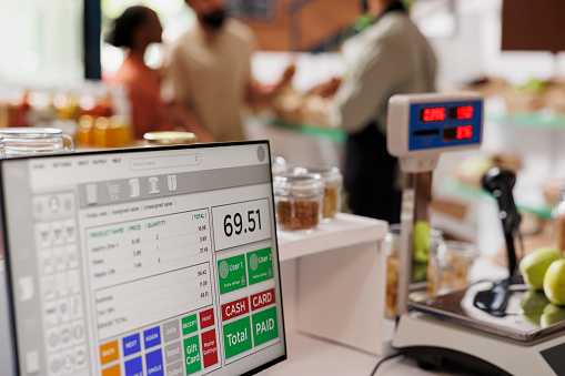 While multiethnic clients are assisted by salesman in the background, apples are placed on digital measuring scale. Photo focus on cashier's desk with weighing equipment and desktop computer.
