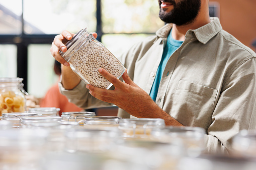 Close up on glass jar filled with locally grown food product held by male consumer in eco friendly convenience store. Detailed image of person grasping plastic free container having organic beans.