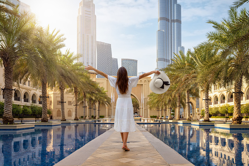A beautiful tourist woman in a white summer dress stands on a public square with palm trees at Downtown Dubai, UAE