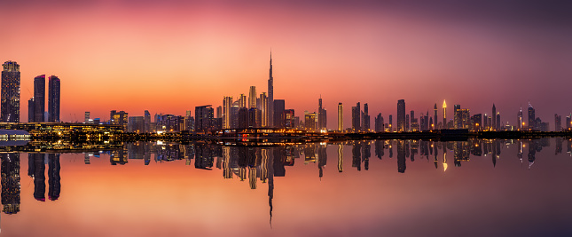 Wide panoramic view of the illuminated skyline of Dubai Business bay with reflections of the modern skyscrapers in the water during dusk time, UAE