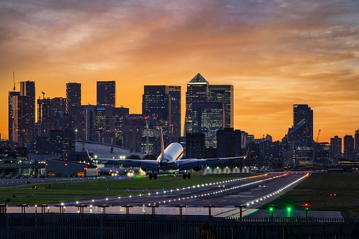 An airplane landing at the illuminated runway of the City Airport in front of the Canary Wharf skyline in London during evening time