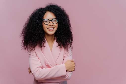 Cheerful young woman with curly hair wearing glasses and a pink blazer
