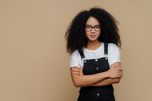 Confident woman with curly hair and glasses in a casual dungaree outfit
