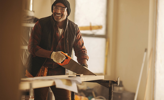 Skilled carpenter cutting wood with a crosscut saw in a well-lit kitchen. He is part of a home renovation project, surrounded by construction equipment and tools for the remodeling work.