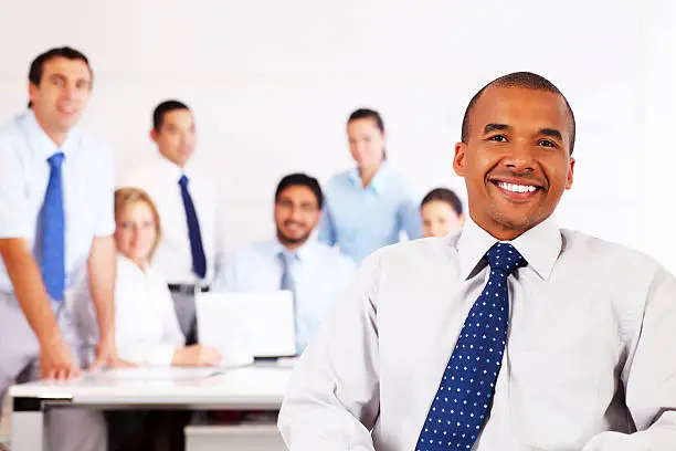 "Smiling African business man sitting and looking at camera,  group of a employees behind him.  White background."