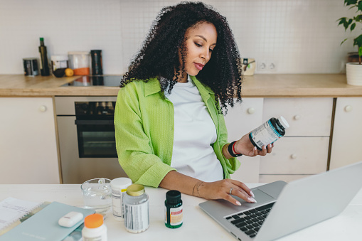 Healthy female with curly hair and dark kin ordering vitamins online, sitting at kitchen table scrolling laptop, filling in form, holding bottle of supplement, examining description and company name