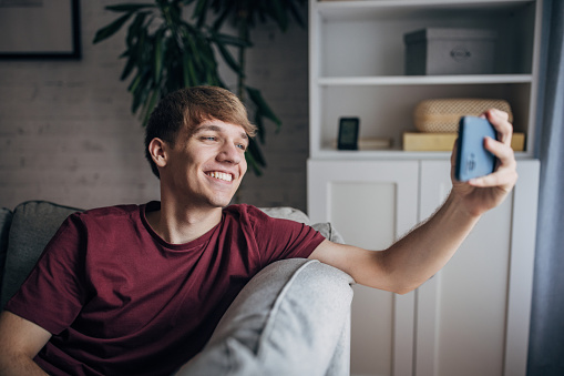 Young man using smart phone to take a selfie while sitting on sofa at home.
