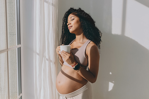Morning coffee and pregnant woman. Happy black female awaiting baby looking through window with cup of hot drink, standing with closed eyes, enjoying moment and her condition of expectant mother