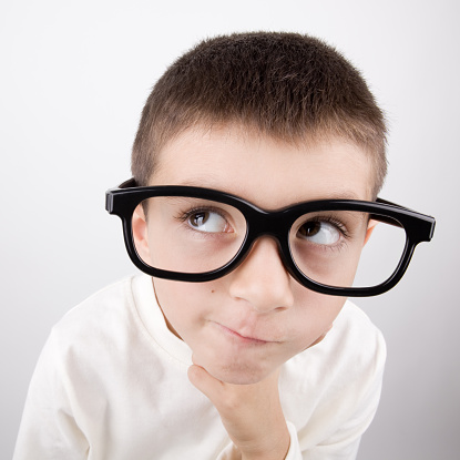 Little boy wearing large horn rimmed glasses and thinking.His hand is under chin and wearing a white sweater.Wide angle view and large glasses are used to have humor effect.The photo was shot with a full frame DSLR camera and cropped to square composition.