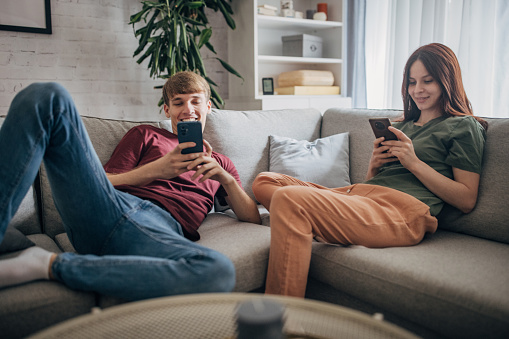 Young couple,  man and woman using smart phones while sitting on sofa at home.