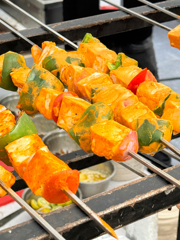 Stock photo showing close-up, elevated view of pieces of marinated paneer and tofu on metal skewers with pieces of red onion, red and green bell peppers lying in rows for barbecuing over hot charcoal briquettes. The buffalo milk curd cheese and bean curd pieces have been marinated in a savoury spicy sauce.