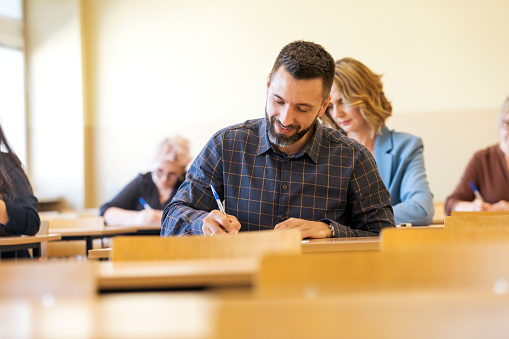 Group of mature adult female and male students sitting in the classroom and writing a test. Focus on mid adult man writing.