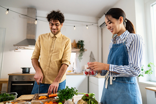 Young Caucasian man cutting and preparing vegetables for weekly usage, while his girlfriend Japanese ethnicity helps him to stage them into glass containers