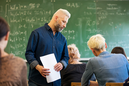 Mature teacher talking to adult students, standing in the classroom with blackboard in the background.