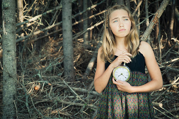 The day is going down in this forest. Somebody is very late. This young girl is getting worried while waiting holding a clock.