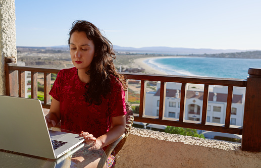 Latin woman sitting on the balcony of her apartment working on the laptop with the ocean and the horizon in the background