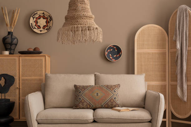istock Warm and cozy living room interior with beige sofa, wooden partition wall, patterned pillow, rattan sideboard, boho lamp, basket on wall and personal accessories. Home decor. Template. 1830409949