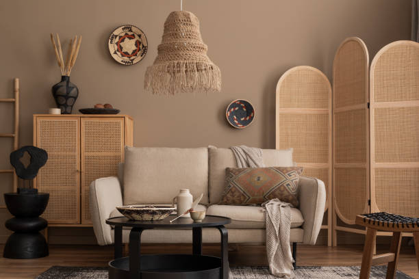 istock Warm and cozy living room interior with beige sofa, wooden partition wall, patterned pillow, rattan sideboard, boho lamp, basket on wall and personal accessories. Home decor. Template. 1830410472