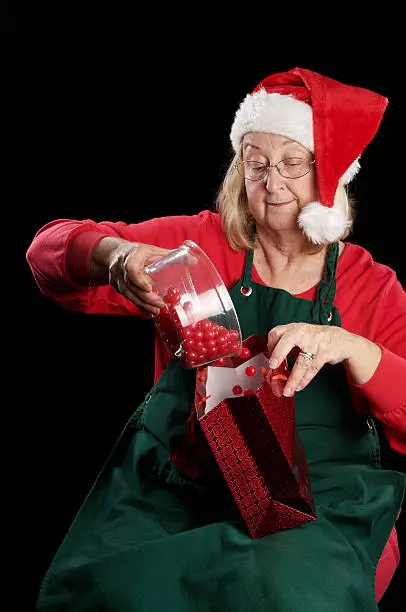 Mrs Claus pours red round candy into red gift bag. Vertical studio shot on black.