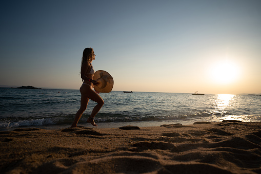 Full length of a carefree woman whit sun hat walking at the beach at sunset