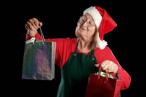 "Mrs Claus holds up silver and red gift bags, smiling. Horizontal studio shot on black."