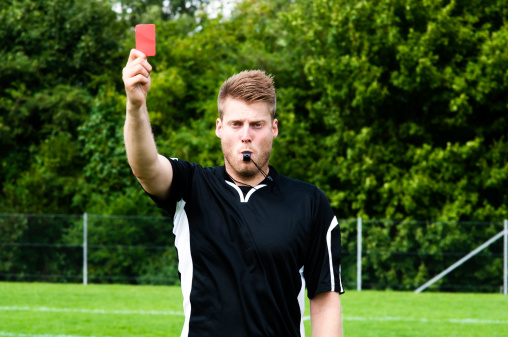 Referee pulling a red card sending a player off the field. The referee is also blowing a whistle.