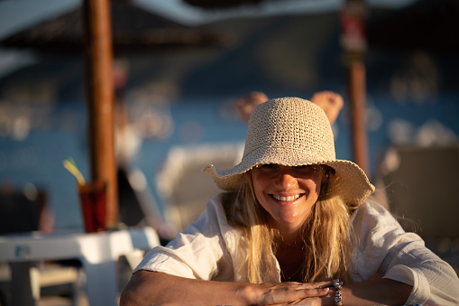 Portrait of a happy blonde woman whit sun hat lying at the beach and looking at camera