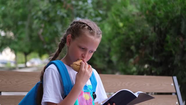 A schoolgirl girl sits on a bench and repeats her lessons. A child with a sports bag in the park eats an apple and studies.
