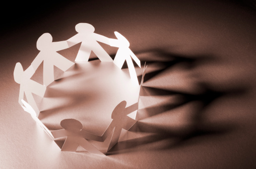 A circle of paper chain people holding hands and facing each other in a circle. There is a side light from the left casting a long shadow of the paper chain to the right. The image has a warm tone. The edges of the frame are very dark in contrast to the plain white paper.
