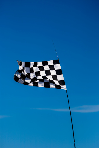 A black and white racing flag is ready to flag the winner against a clear blue sky. Vertical composition. Adobge RGB 1998 profile.