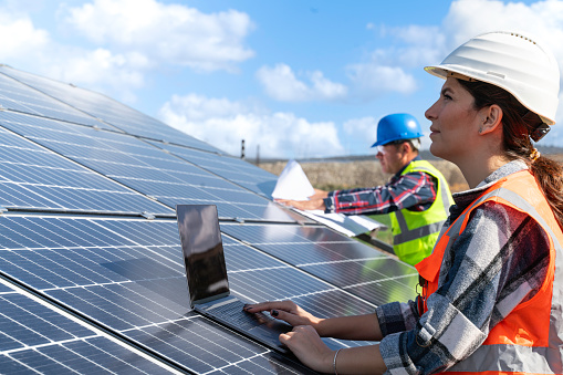 Young engineer inspect installation of solar panels installed check  with blueprint on the field, with recording inspection data on pc. Two technicians in distance discussing between long rows of photovoltaic panels. Solar panel installer installing solar panels with a big copy space