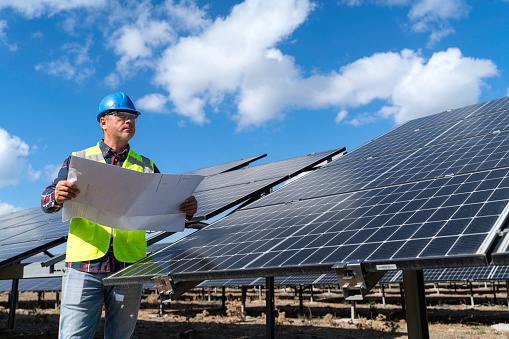 Young engineer inspect installation of solar panels installed check  with blueprint on the field, with recording inspection data on pc. Technicians in distance discussing between long rows of photovoltaic panels. Solar panel installer installing solar panels with a big copy space