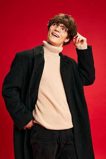 Portrait of young happy and smiling man, student in knitted beige sweater and coat thoughtfully scratches his head against red studio background. Concept of human emotions, beauty, fashion and style.