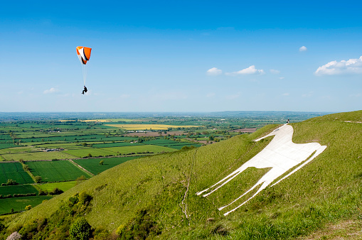 A wide angle view of a paraglider airborne over the famous landmark of the Westbury White Horse hill figure in Wiltshire. Cut into the hillside in 1778, this hill carving replaced an older horse which was possibly cut to commemorate King Alfred's nearby victory over the Vikings. Image taken on a windy summer day in late afternoon with a ProPhoto RGB profile for maximum color fidelity and gamut.