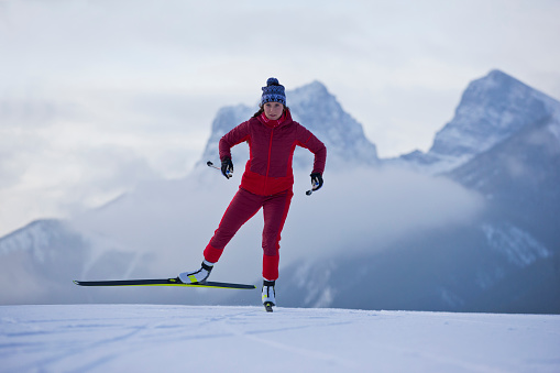 A female cross-country skier goes downhill at the Canmore Nordic Centre Provincial Park in Alberta, Canada. She is doing the skate skiing, or freestyle, technique and is using skate skiing equipment. In the background are two of the Three Sisters mountain peaks.