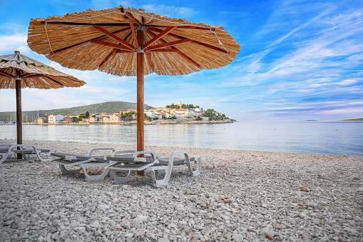Straw umbrellas with sunbeds on the beach. Straw umbrellas and stacked deck chairs on the beach at sunset or sunrise. Primosten, Croatia.