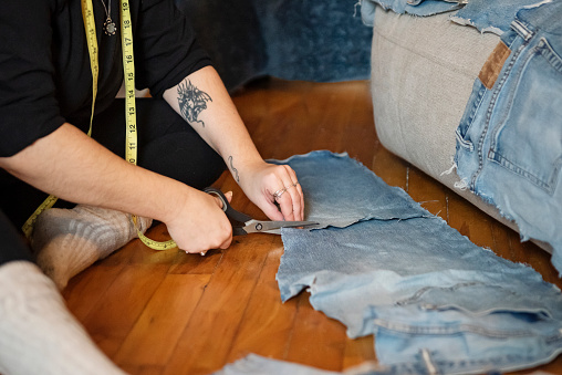 Close-up on woman’s hand upcycling old jeans to cover worn out sofa. She is a body positive member of the LGBTQ community. She is dressed in casual black leggings and top. Obscured face. Horizontal close-up indoors shot with copy space. This was taken in Montreal, Quebec, Canada.