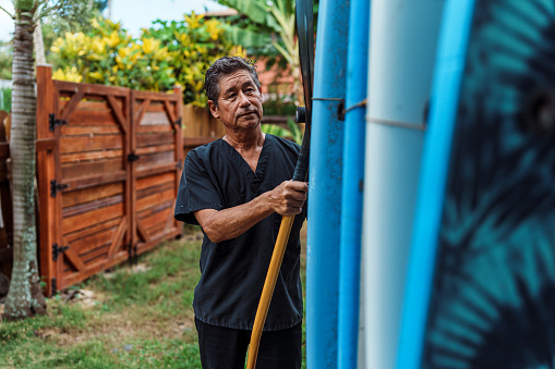 A vibrant mature adult multiracial man of Hawaiian and Finnish descent who is wearing scrubs stands outside his house and chooses a surfboard to load into his car before heading to work in the morning.