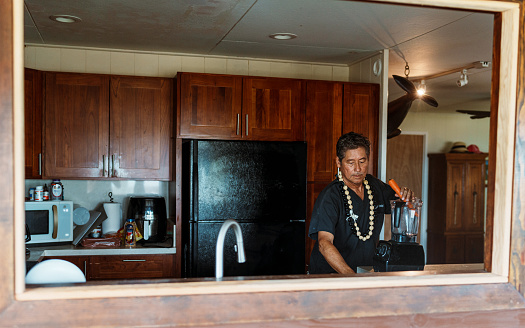 A mature adult multiracial man of Hawaiian and Finnish descent who is wearing scrubs uses a blender to prepare a healthy smoothie in the kitchen of his home.