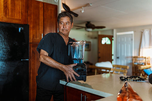 A mature adult multiracial man of Hawaiian and Finnish descent who just arrived back home from work and is wearing scrubs picks a blender up off the kitchen counter to put it away.