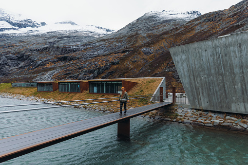 Side View of female in yellow hat and jacket walking at beautiful viewpoint on top of the mountain range with background view of snowcapped peaks in Trollstigen Western Norway, Scandinavia