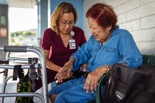 A caring Eurasian mature adult occupational therapist places a transfer gait belt on a female elderly patient of Asian descent that she is visiting outside the female patient's apartment.