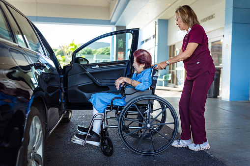 A multiracial female healthcare worker who is wearing scrubs pushes an elderly woman of Asian descent in a wheelchair toward a parked car outside a medical facility.