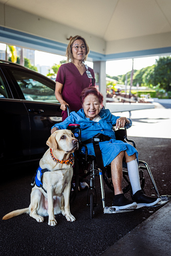 A cheerful elderly woman of Asian descent who is using a wheelchair poses with her emotional support service dog outside her apartment building with her home caregiver.