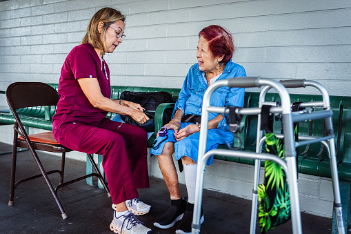 An occupational therapist sits outside on a folding chair and uses a finger pulse oximeter to check vitals on her patient, a smiling elderly woman of Asian descent.