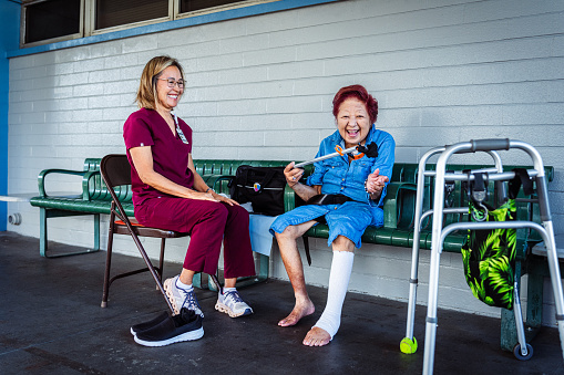 A geriatric woman of Asian descent smiles with excitement while holding a grabbing aid her occupational therapist taught her to use to remover her socks and shoes.